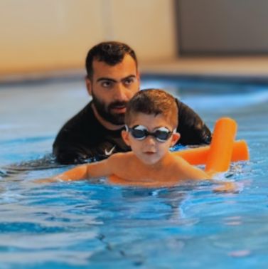 ילד עם משקפי שחייה שוחה בעזרת נודל כתום, בעוד מדריך עומד מאחוריו בתוך הבריכה ומלווה אותו מקרוב.