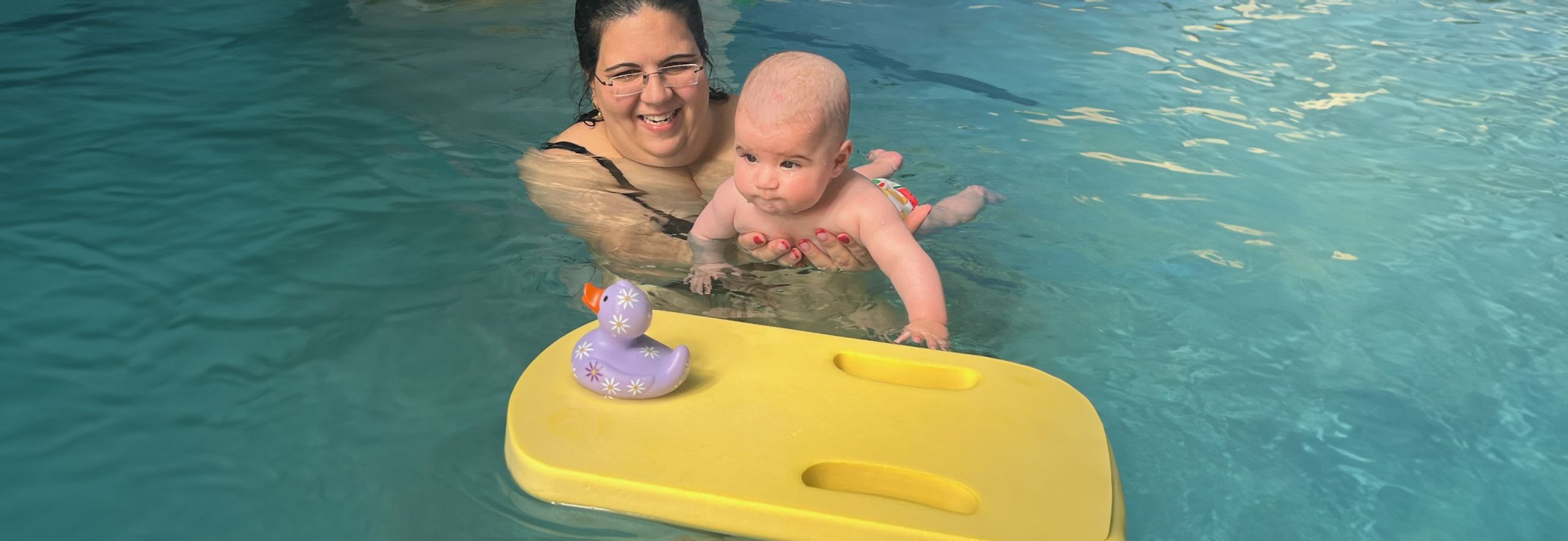 אם מחזיקה תינוק במים בזמן פעילות בבריכה, והתינוק נשען על מצוף צהוב שעליו מונח ברווז צעצוע סגול