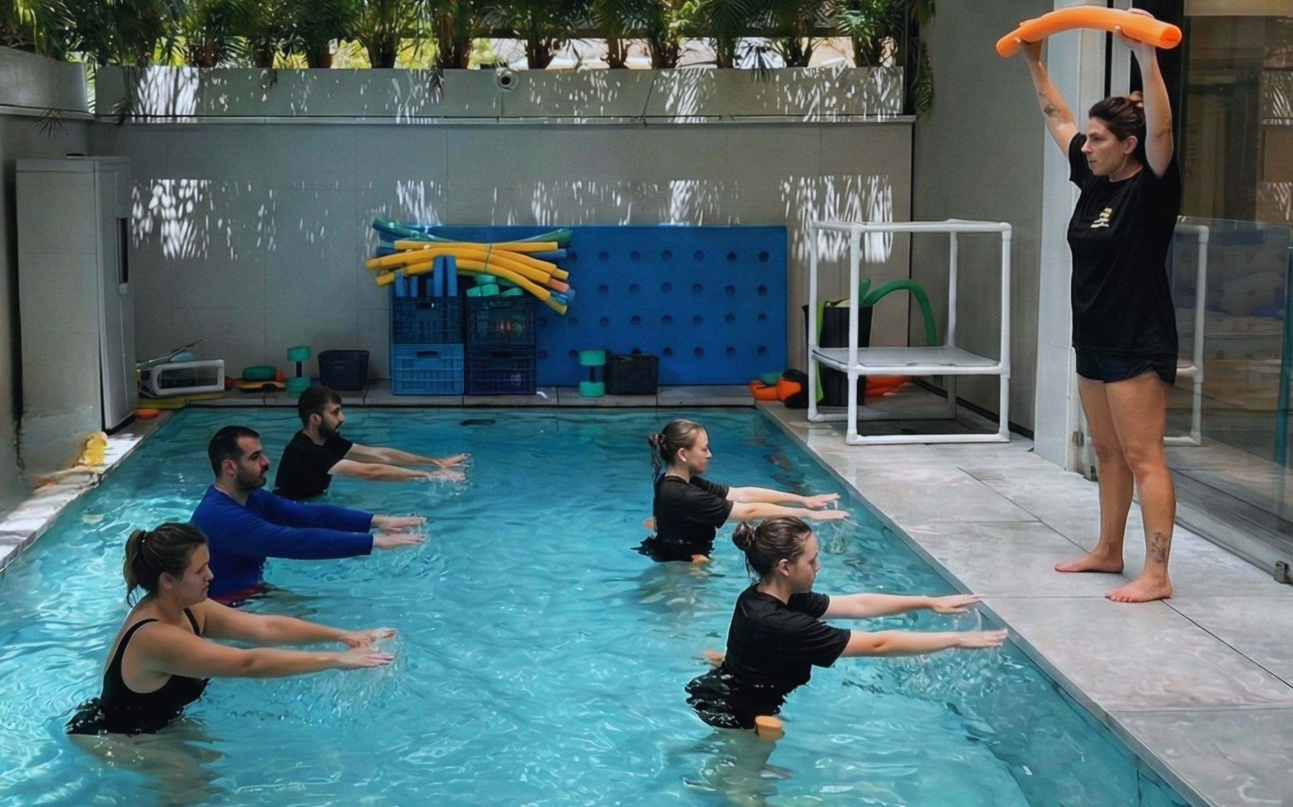 מבוגרים עומדים בשורה בתוך בריכה בשיעור פעילות במים, מותחים את הידיים קדימה לפי הנחיית מדריכה שמחזיקה נודל כתום מעל ראשה